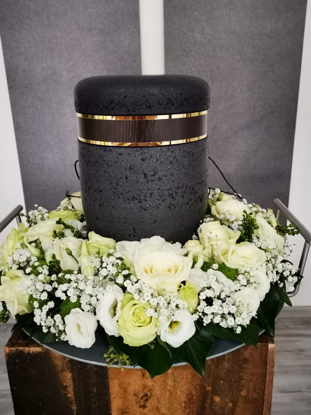 Schwarze, strukturierte Urne mit Goldrand, umrahmt von einem Kranz aus weißen Rosen und Schleierkraut auf einem Holzsockel.