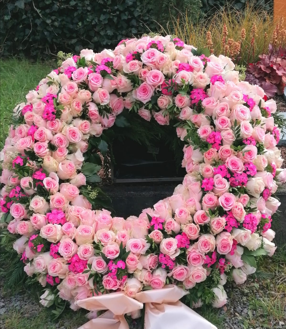 Großer, stehender Trauerkranz aus hellrosa Rosen und kräftig pinkfarbenen Bartnelken im Freien auf Rasen.
