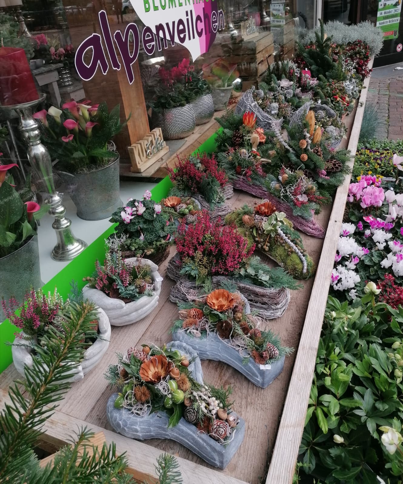 Ausstellung verschiedener Blumengestecke, saisonaler Topfpflanzen und Grabschmuck auf Holztischen vor einem Blumenladen.